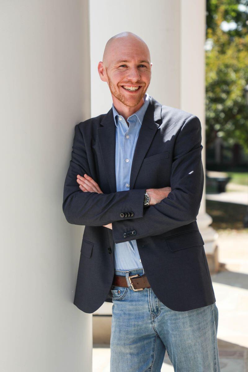 Photo of a white male in a suit jacket and jeans leaning on a white column outside.