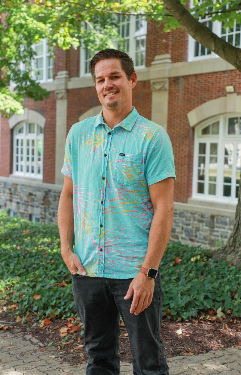 Photo of white male standing in front of a brick building wearing a blue patterned shirt.