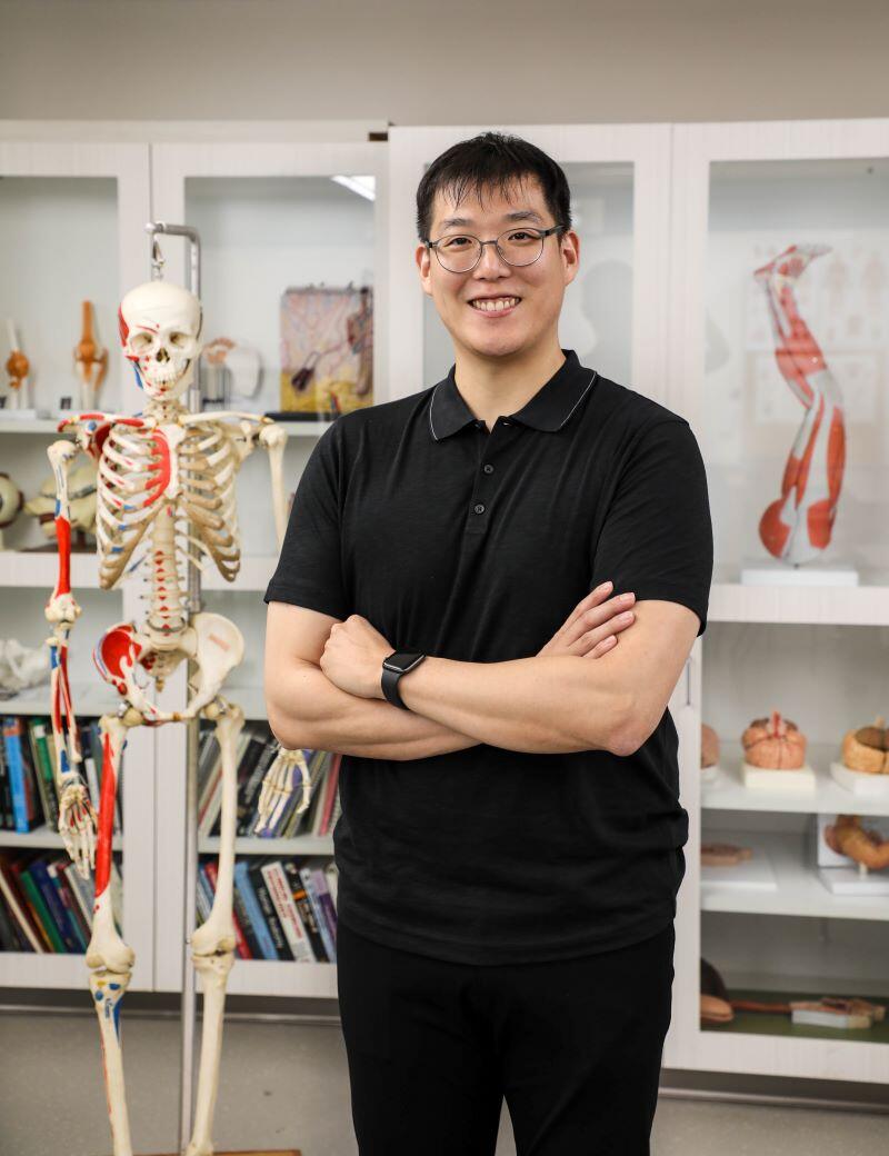 Photo of an Asian male in a black shirt standing in an anatomy lab with a model skeleton to his right.