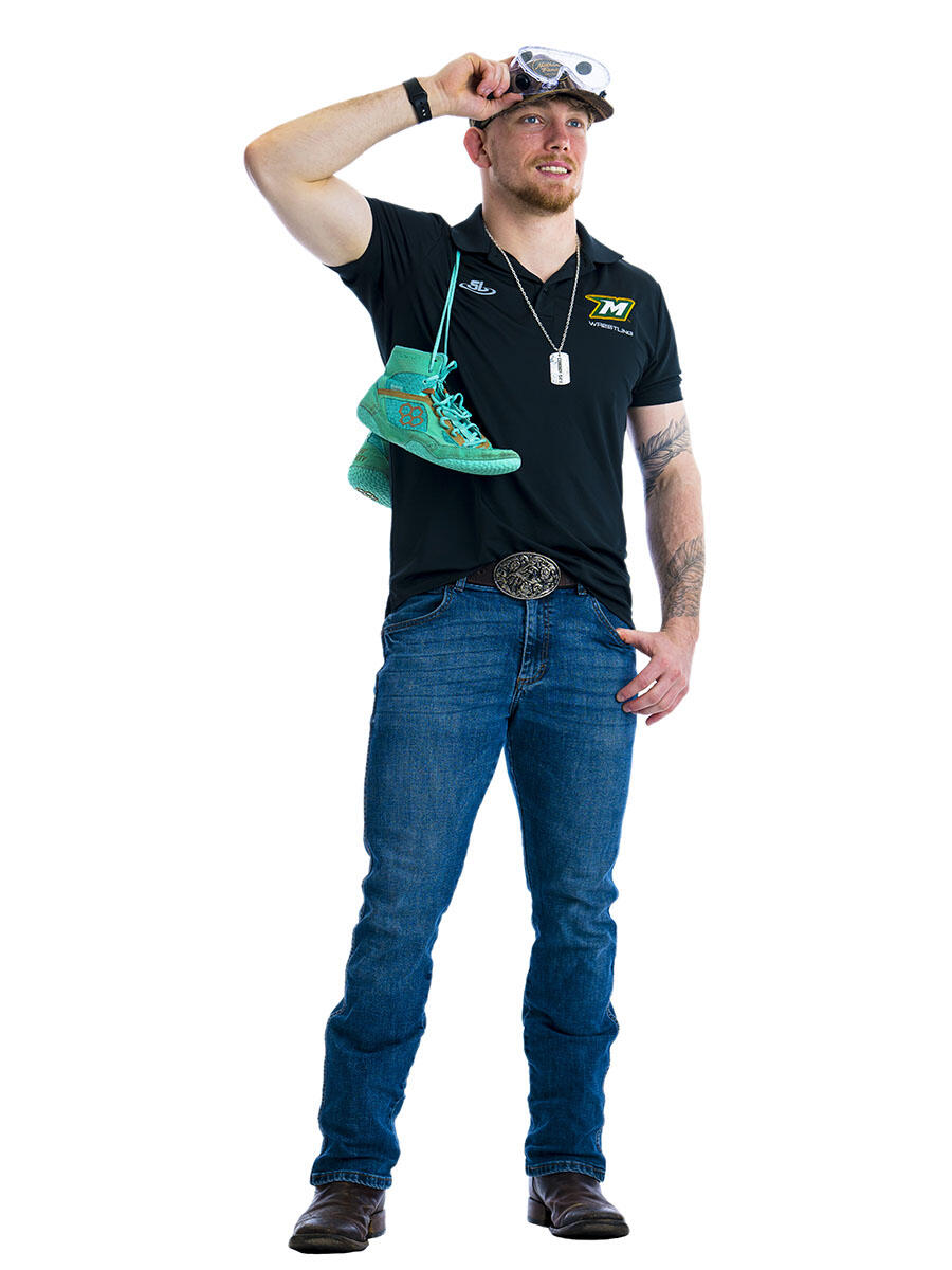 A student stands in front of white background with one hand on a pair of science goggles on his head and a pair of wrestling shoes draped over his shoulder.