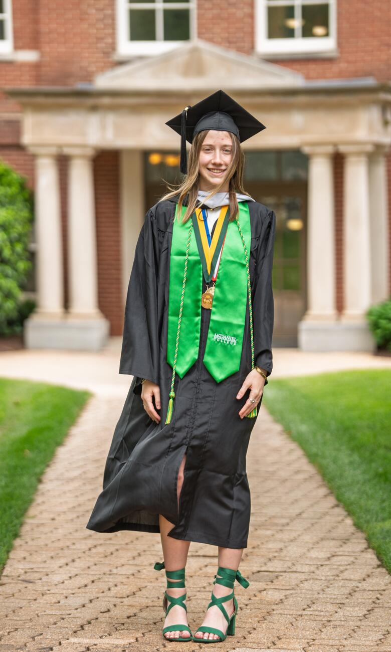 Lorelei McElroy stands in front of a red brick building at McDaniel while wearing her graduation robe and cap with a green stole.