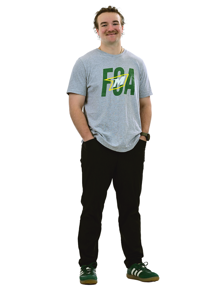 A male student wearing a gray McDaniel shirt and black pants stands with his hands in his pockets.