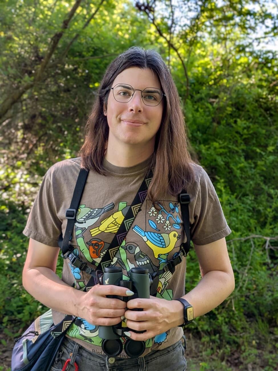 person with brown hair stands in front of trees
