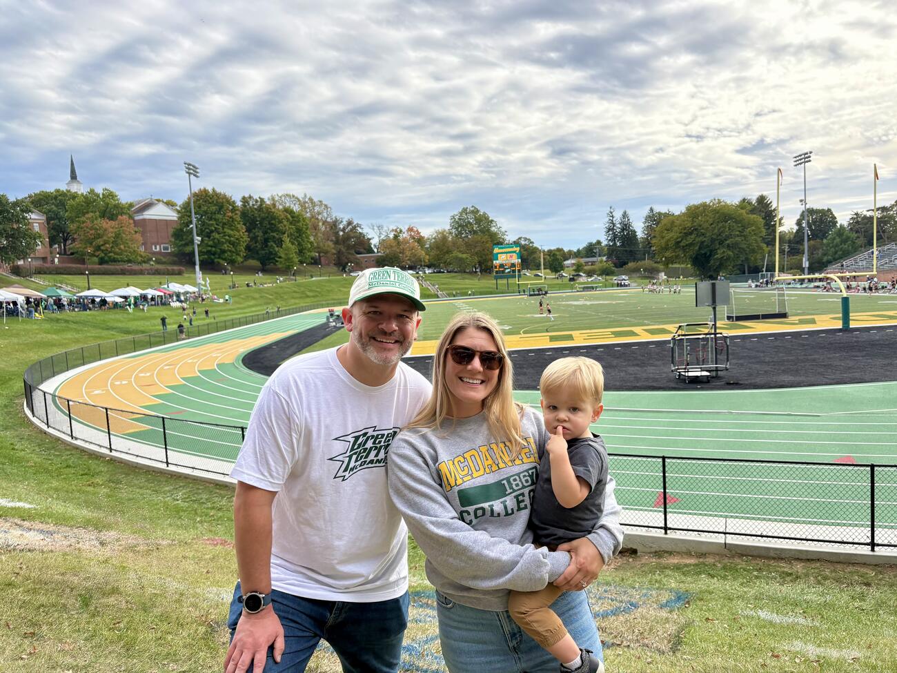 A man and woman wear McDaniel gear and hold a toddler in front of the McDaniel football stadium.