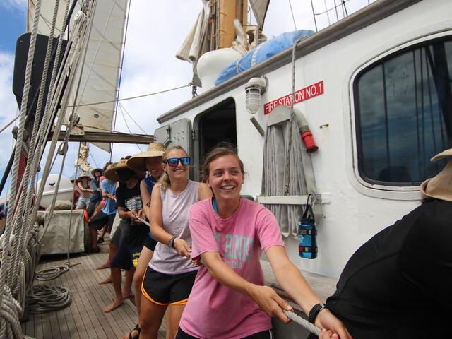 Riley Palmer '18, in a pink shirt, helps raise the main sail on board the SEA Semester ocean research vessel, Robert C. Seamans.