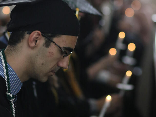 Graduates in cap and gown seated holding candles during ceremony.