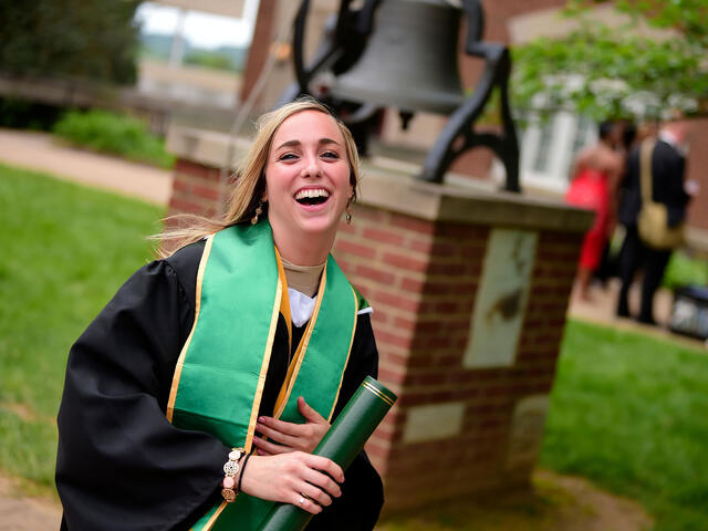 Graduate in cap and gown standing near the Bell.
