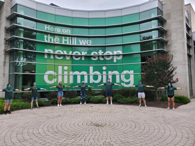 Seven students stand six feet apart in front of McDaniel College Gill Center sign that reads "Here on the Hill we never stop climbing".