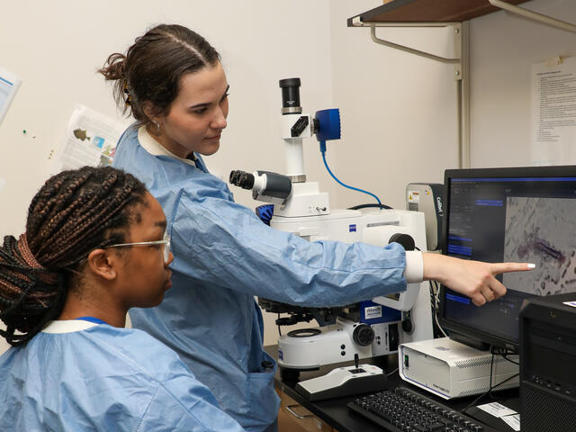 Two students in lab coats look at a microscope.