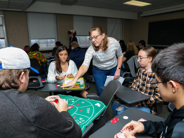 A group of students plays Tripoley in a classroom. The professor points at the board.