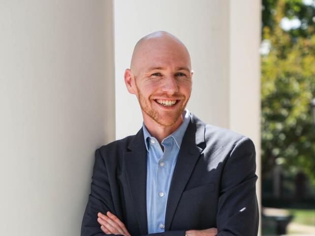 Photo of a white male in a suit jacket and jeans leaning on a white column outside.