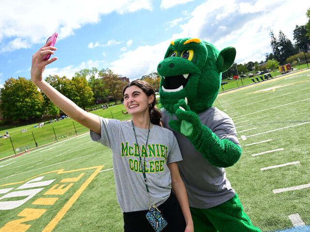 A female student in a McDaniel College t-shirt poses with the Green Terror mascot while taking a photo on her phone.