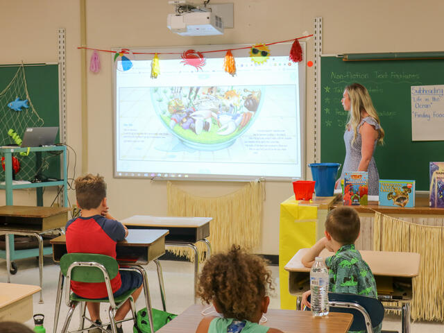 A female teacher looks at a projector image while standing at the front of a classroom with young children at desks.