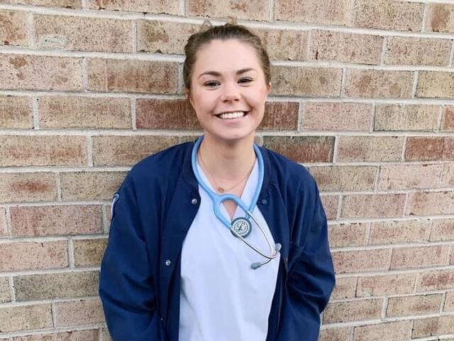 A woman in nursing scrubs smiles while leaning against a brick wall.