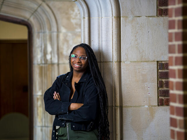 A student stands with arms crossed in a hallways with a stone arch.