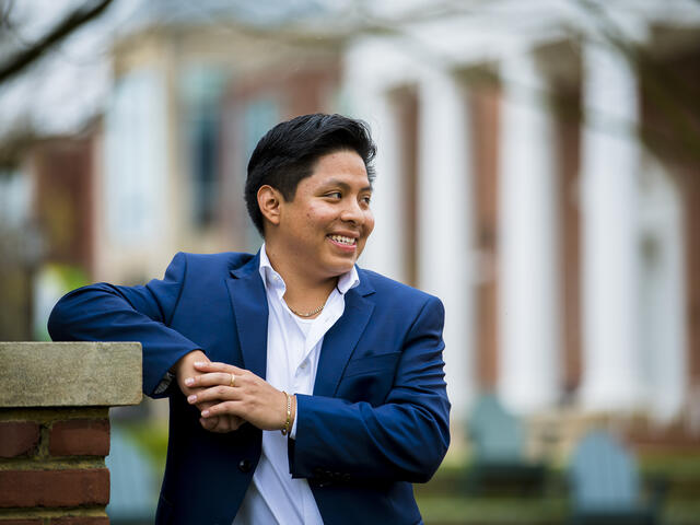 A student wearing a suit leans his elbow on a pedestal while smiling off camera.