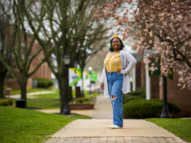 A student in a yellow shirt and grey cardigan stands outside in front of a row of cherry blossom trees.