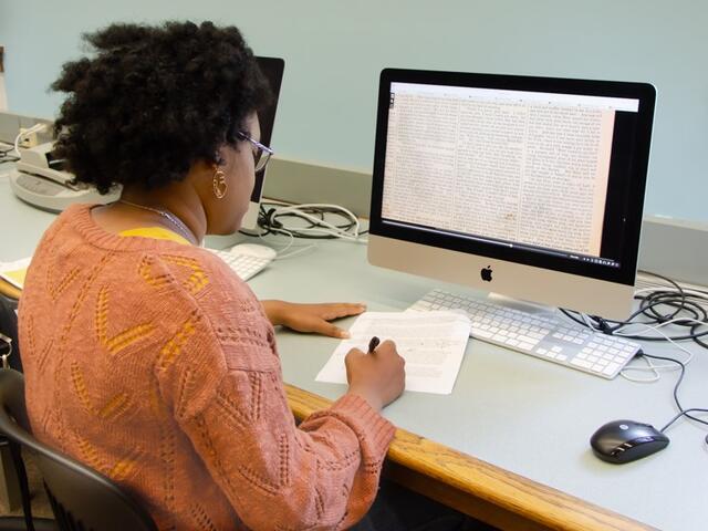 A student in an orange cardigan writes on a piece of paper while looking at an image of a newspaper story on a computer screen.