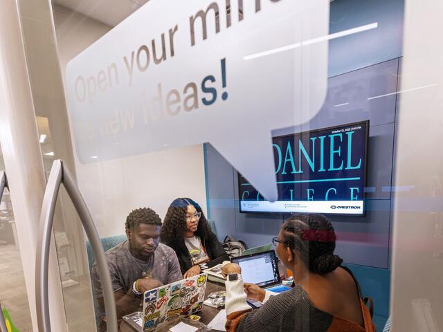 Three students sit together in a room with glass walls, each on their laptops and talking. A decal on the glass reads "Open your mind to new ideas!"