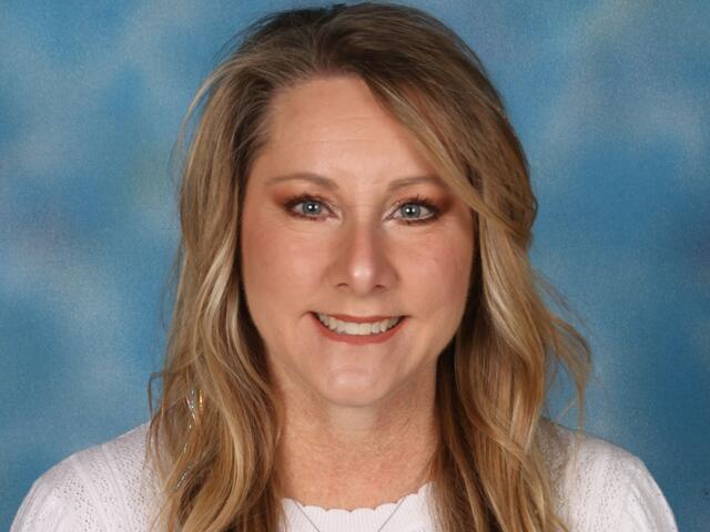 Kristina Stacey, a woman with long, blonde hair, wears a white sweater and smiles in front of a blue backdrop.