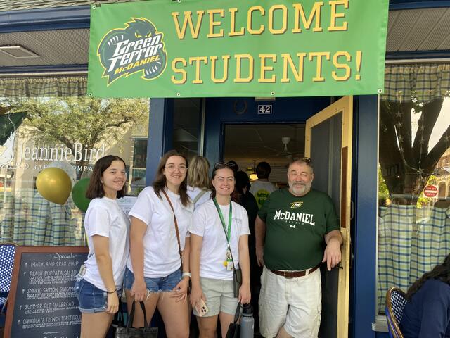 Bernie Vogel of JeannieBird Baking Company with students under a sign saying "Welcome Students"
