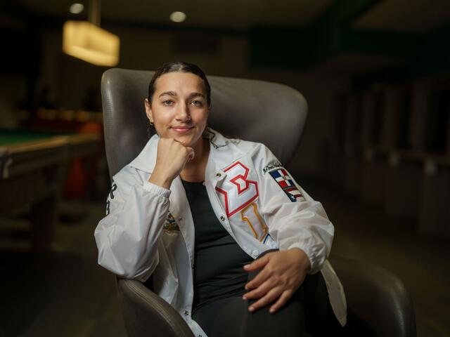Katherine Taveras sits in a chair while leaning her head on her hands. She wears a white jacket with the logo for her sorority, Hermandad de Sigma Iota Alpha.