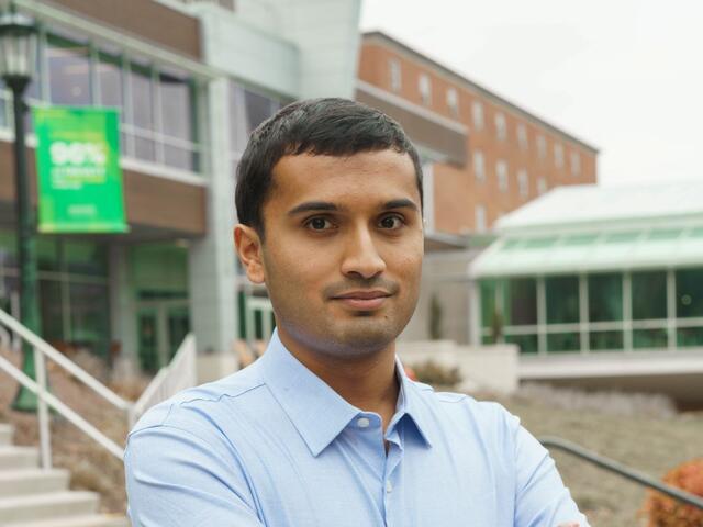 Nikhil Niyogi stands outside of the student center with his arms crossed, wearing a blue button-down shirt, a belt, and tan pants.