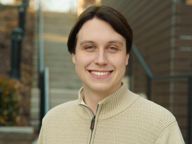 Headshot of Paul Steinert standing outside in front of a staircase, wearing a tan sweater.