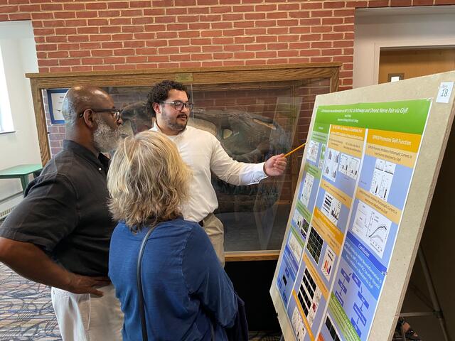 A male student points to a research poster board while two people listen.