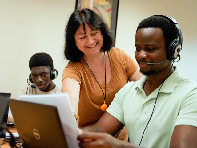 A McDaniel French professor in an orange shirt leans between two students wearing headsets who are seated at computers.