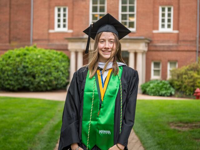 Lorelei McElroy stands in front of a red brick building at McDaniel while wearing her graduation robe and cap with a green stole.