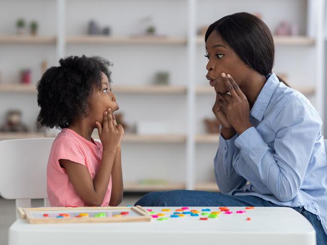 A young woman sits next to a small child at a table. They both hold their hands up to their face while shaping a sound with their mouths.