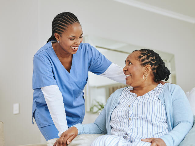 A young African American woman in scrubs leans over a senior African American woman patient while they both smile.