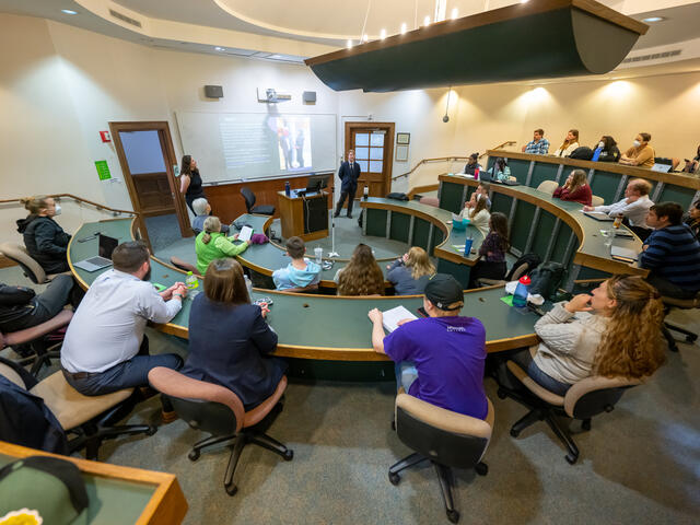 students sit at desks in an auditorium style classroom with a professor standing in the front