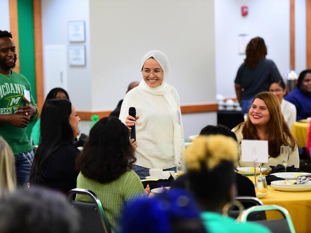 A smiling student holds a microphone while engaging in conversation with classmates at round tables during the annual Scholarship Luncheon