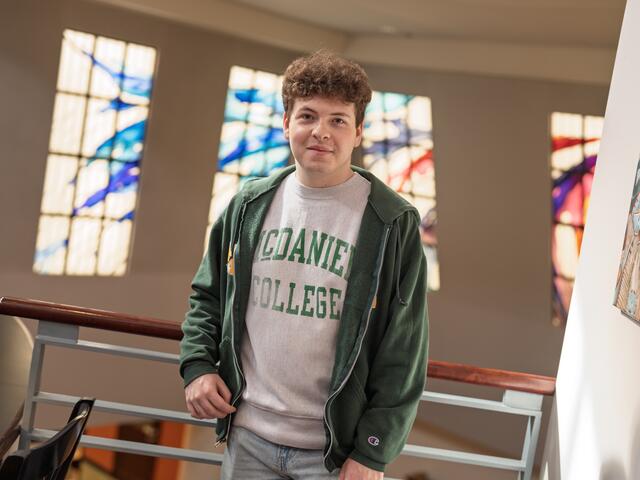 A male student in a McDaniel College shirt stands in front of stained glass windows.