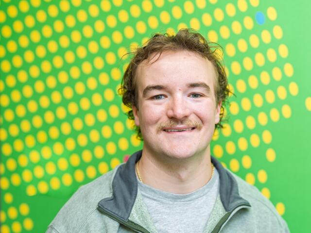 A male student smiles in front of a green patterned wall.
