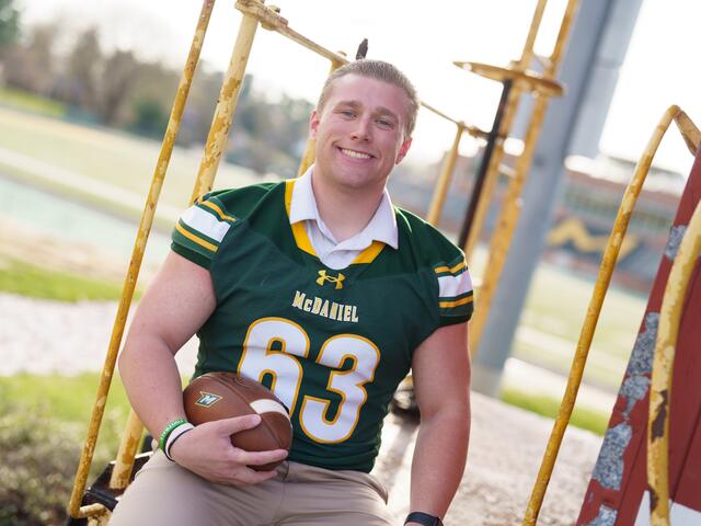 A male student wearing a football jersey smiles and holds a football.