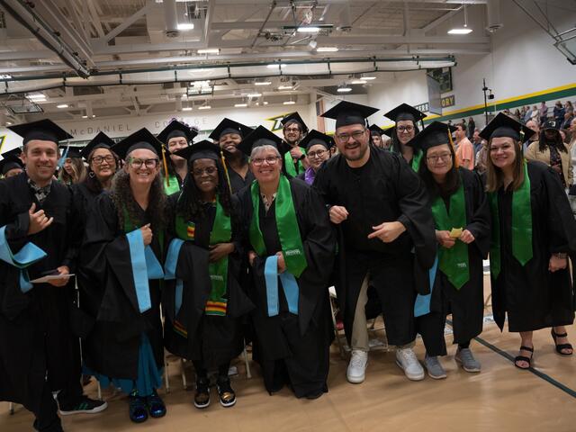 A large group of graduates in caps, gowns, and green McDaniel College stoles pose together smiling in the gymnasium before the graduate commencement ceremony.
