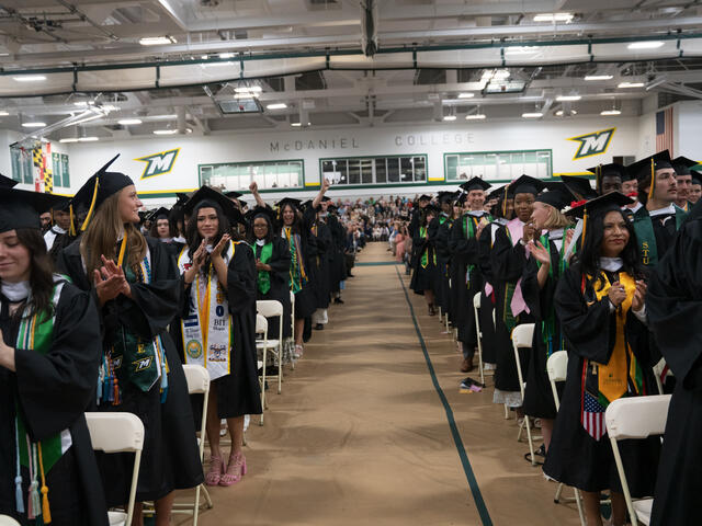 students in graduation regalia stand in rows with an aisle down the middle