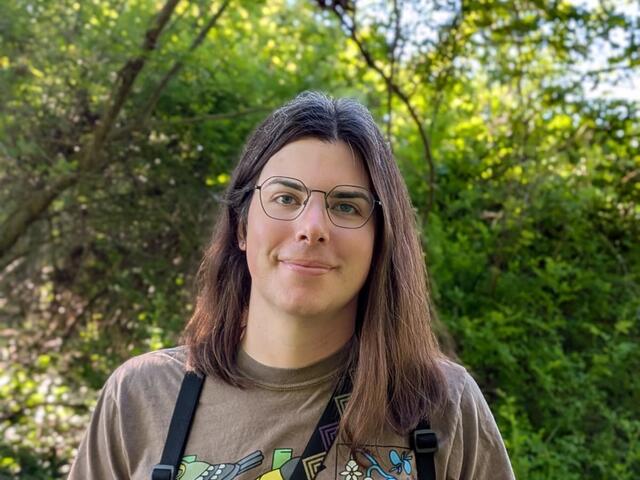 person with brown hair stands in front of trees