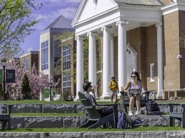 Two students sitting in Adirondack chairs on a terraced lawn, talking and using a laptop, with a columned campus building and blooming spring trees visible in the background.