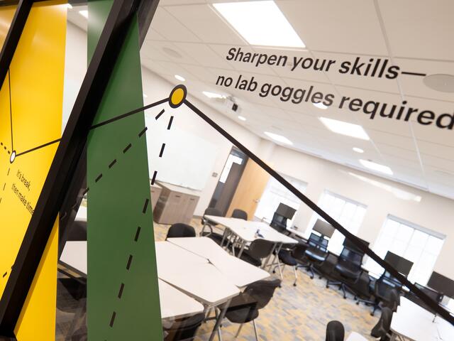 Close-up angled view through a glass window decal at the STEM Center, showing yellow and green diagonal stripes with geometric line graphics and text reading "Sharpen your skills—no lab goggles required," with a classroom of tables, chairs, and monitors visible beyond the glass.
