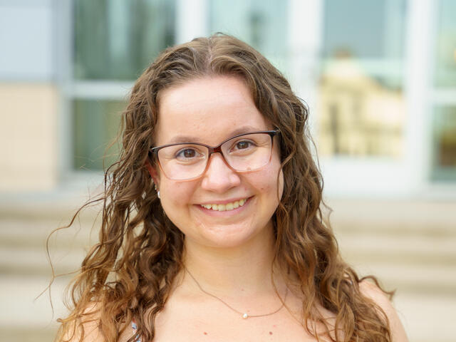A young woman with long brown hair and glasses smiles in a floral dress.