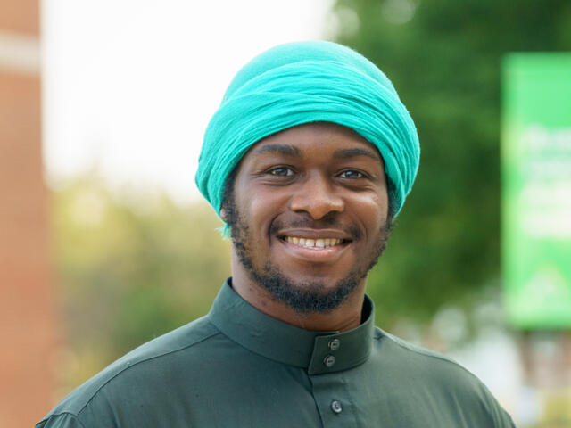 A young man smiles in a turquoise turban and a dark green shirt.