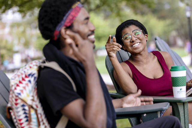 Students laughing and in conversation sitting in outdoor chairs.