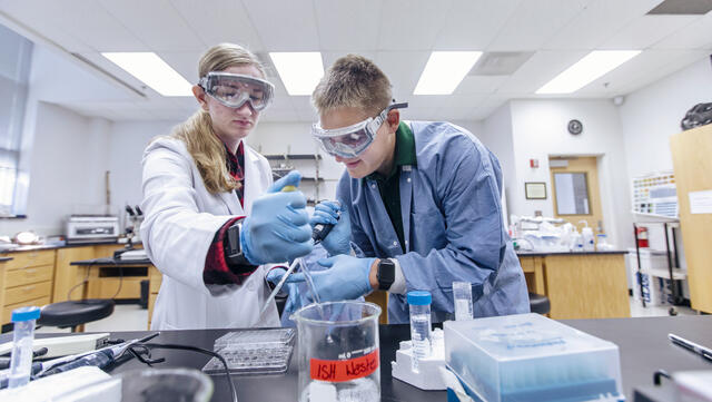 Two students in lab gear create a chemistry solution.