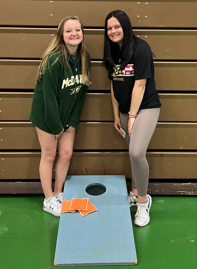 Two students pose by a cornhole board.