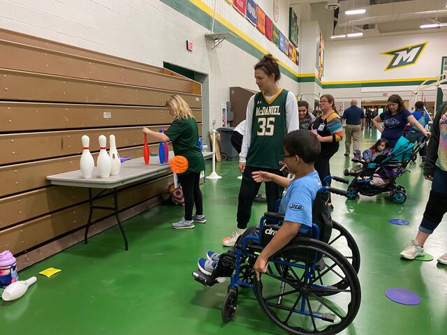 A student helps a child bowl by tossing a ball at a set of bowling pins.