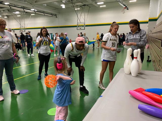 A student helps a child bowl by tossing a ball at a set of bowling pins.